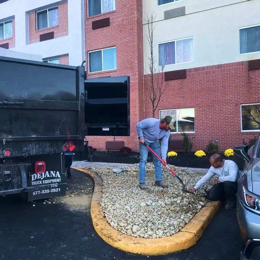Two men are working in a landscaped area outside a multi-story brick building.